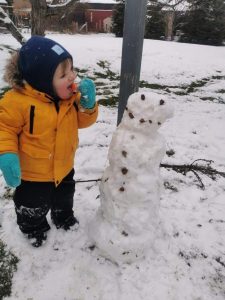 A little girl eats a carrot from a snowman's nose. 