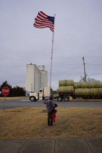 Boy waves flag at passing truck with hay bales.